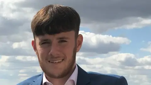 Abrahams Family A close up photo of a man with a brown beard, short brown hair wearing a pink shirt and a blue blazer.