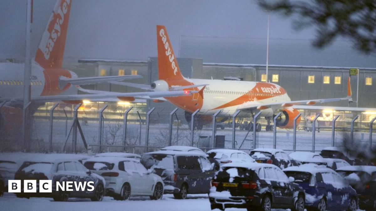 EasyJet planes are grounded due to snow and ice on the runway at Liverpool John Lennon Airport. There are snow-covered cars parked outside the airport's fence.