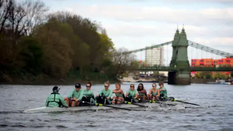 John Walton/PA Wire An eight-person racing rowing boat on the River Thames in London. The rowers are wearing the pale "Cambridge Blue" sports kit and are facing towards the camera. A coxswain with "Cambridge" written across their back is seated closest to the camera, but is facing away from us. The oars are all in the water as the rowers are pulling through a stroke. Behind the boat in the far distance is an imposing road bridge. There are trees and bushes on the left of the image, where the river bank meets the water.