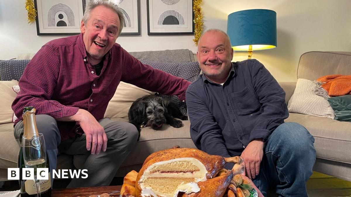 Paul Whitehouse and Bob Mortimer are grinning as they sit in a lounge. The roast turkey cake is on a table in front of them and it has been carved to reveal a Victoria sponge cake. There is also a bottle of champagne with a glass. There is a small black dog lying on the sofa next to Whitehouse and tinsel above the picture behind them.