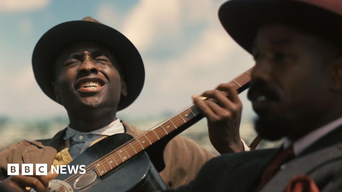 A still from Sinners showing Miles Caton as Sammie playing a guitar and singing and Michael B Jordan in the foreground looking ahead as he drives a car