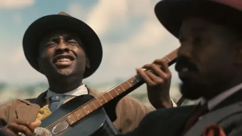 Warner Bros A still from Sinners showing Miles Caton as Sammie playing a guitar and singing and Michael B Jordan in the foreground looking ahead as he drives a car
