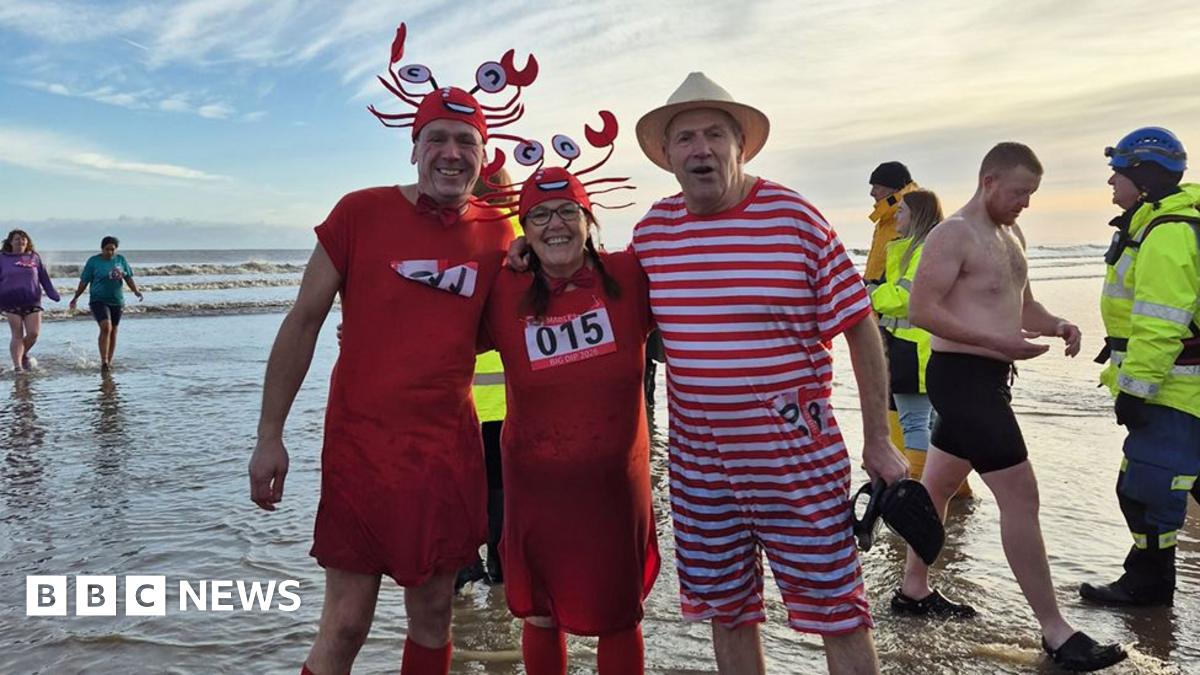 Three swimmers pose for the camera - a man and a women are wearing red crab outfits with one man wearing a red and white striped Victorian bathing costume with hat