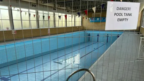 Ann Gannon / BBC An indoor swimming pool stands fully drained, with a metal barrier placed around the empty tank. A sign on the barrier reads “Danger! Pool tank empty”. Sunlight enters through the long row of windows, and colourful lane flags hang above. The tiled floor and exposed pool fittings are visible