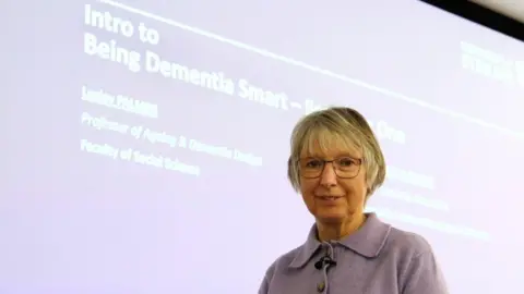 Joan Lyon, a 75-year-old woman, with grey bobbed hair and glasses, wearing a lilac cardigan, stands in front of a big screen with a slide that says Intro to Being Dementia Smart.