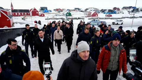 Reuters Danish PM Mette Frederiksen, in the centre of the picture wearing a light-brown puffer jacket, walks with the Greenland prime minister and a large crowd towards the camera on an ice-covered street in Nuuk with houses and a church in the background.