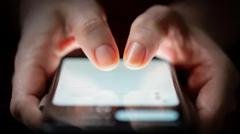 Getty Images Two hands holding a smartphone. The screen of the phone is lit up and the background is dark. Both thumbs are poised to type in a number.