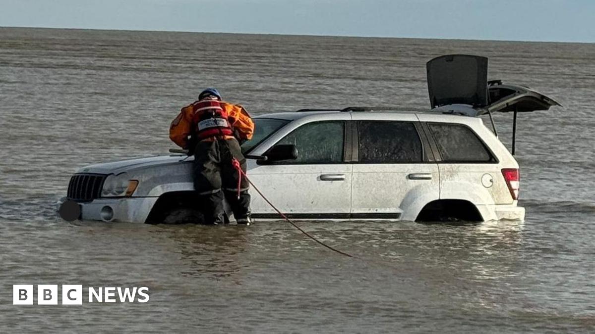 A person in an orange and black jumpsuit leaning on a silver car that has water up to the top of its wheels.