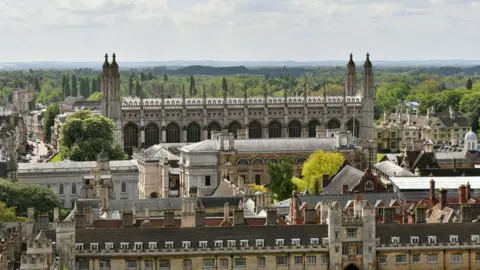 Joe Giddens/PA Wire A view over the rooftops of the city of Cambridge. Prominent in the middle of the image are the roof and spires of the chapel at King's College, Cambridge, which rises above the surrounding buildings. It is rectangular and we are looking towards one of the chapel's longer sides. From each of its four corners rise tall spires and along the side of the chapel are a series of arched windows. There are other, handsome older buildings in the foreground, while in the distance are green trees and the horizon, as the city gives way to the south Cambridgeshire countryside. 