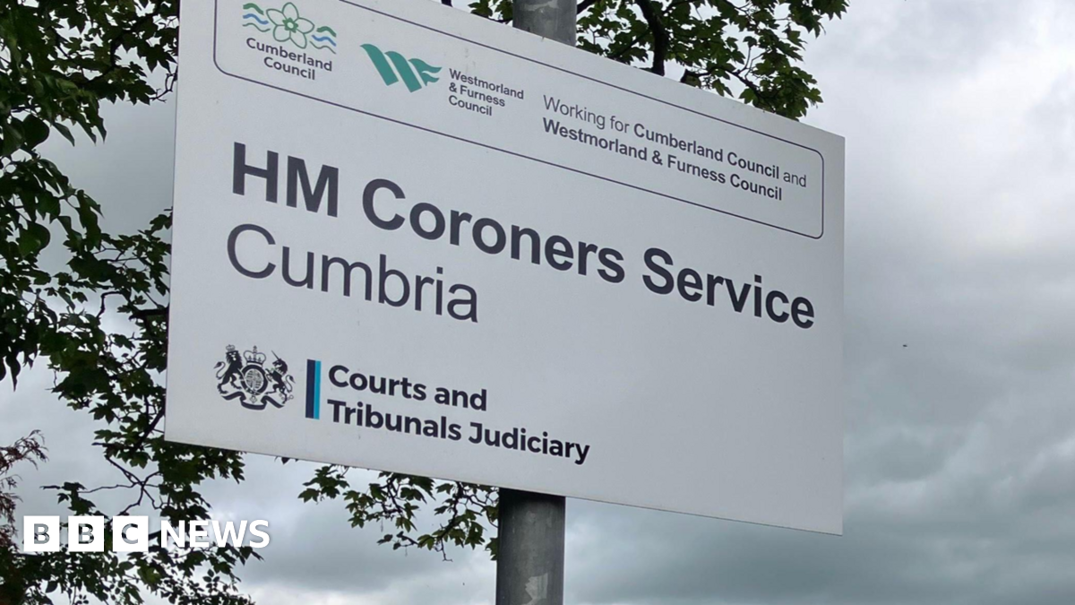 The sign at the entrance to the coroner's court in Cockermouth. It is a white sign on a silver metal pole  with various logos and names of government organisations, with the most prominent words across the middle reading HM Coroners Service Cumbria. The gable end of a brick two-storey building with a pointed tiled roof stands at right angles to a single storey flat-roofed extension which appears to house the main entrance. Behind the smaller building is a Sainsbury's shop front. Cars are parked outside the coroners office. On the opposite side of a narrow street, with no pavement, to the right of the building building are much older two and three storey stone buildings in a terrace.