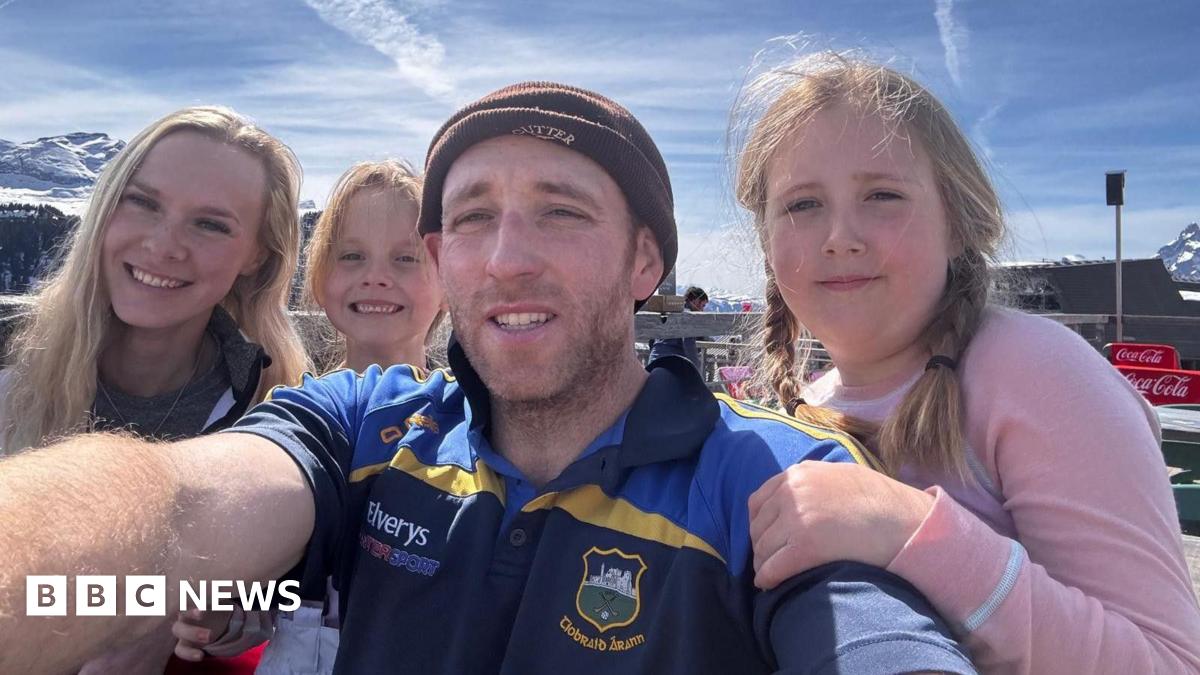 James holds out the camera for a selfie with his family of two girls and his partner Ellie. They appear to be in a ski resort as there are snowy mountain tops behind. He wears short sleeved T-shirt and beany hat.