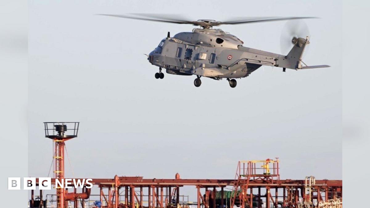 A grey helicopter seen above an orange and blue oil tanker