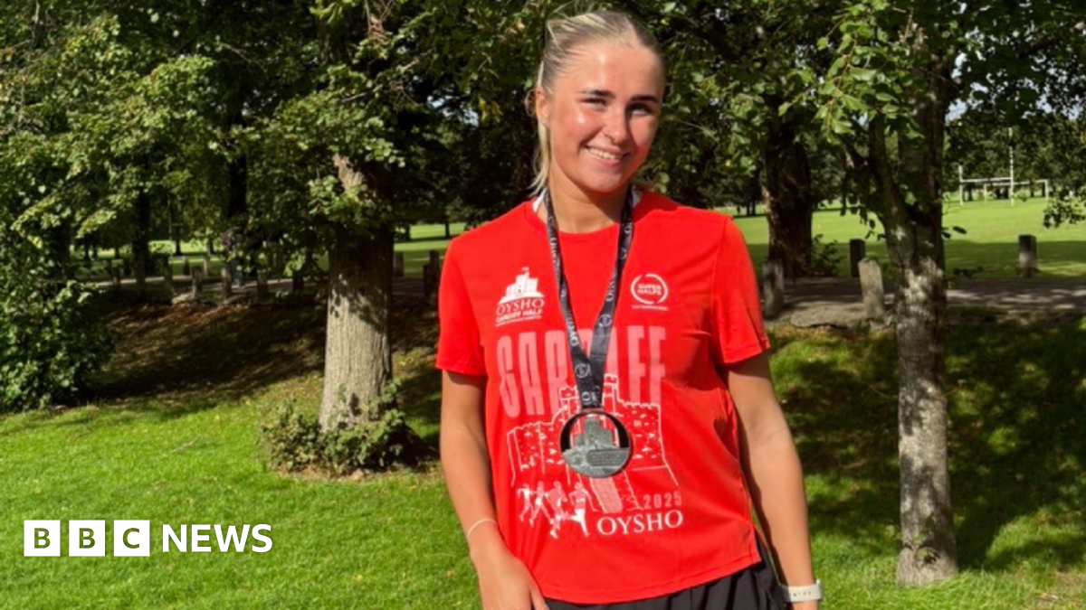 Leah smiling looking at the camera. She is stood in front of trees and greenery and wears a red Cardiff half marathon finishers t-shirt, a black medal and black running shorts. Leah has blonde hair in a high ponytail.