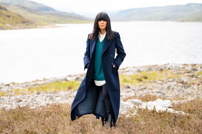 Claudia Winkleman stood in the middle of a marsh, posing as she looks ahead.She is wearing a blue outfit paired with a long blue trenchcoat.