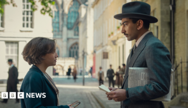 A young woman stands in profile on the left, wearing a blue coat, looking up at a man wearing a grey suit and hat, with a newspaper under his arm. They are stood on a regency-era-style street.