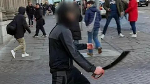 Alan Simpson A teenager wearing all black clothing carrying a large bladed weapon with an orange handle.
