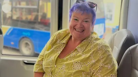 Carolyn Harris An overweight woman on a bus, with purple hair and a mustard-coloured dress.