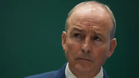 PA Media A close up picture Micheál Martin, a man with thin grey hair, wearing a suit and standing up in front of a green background.