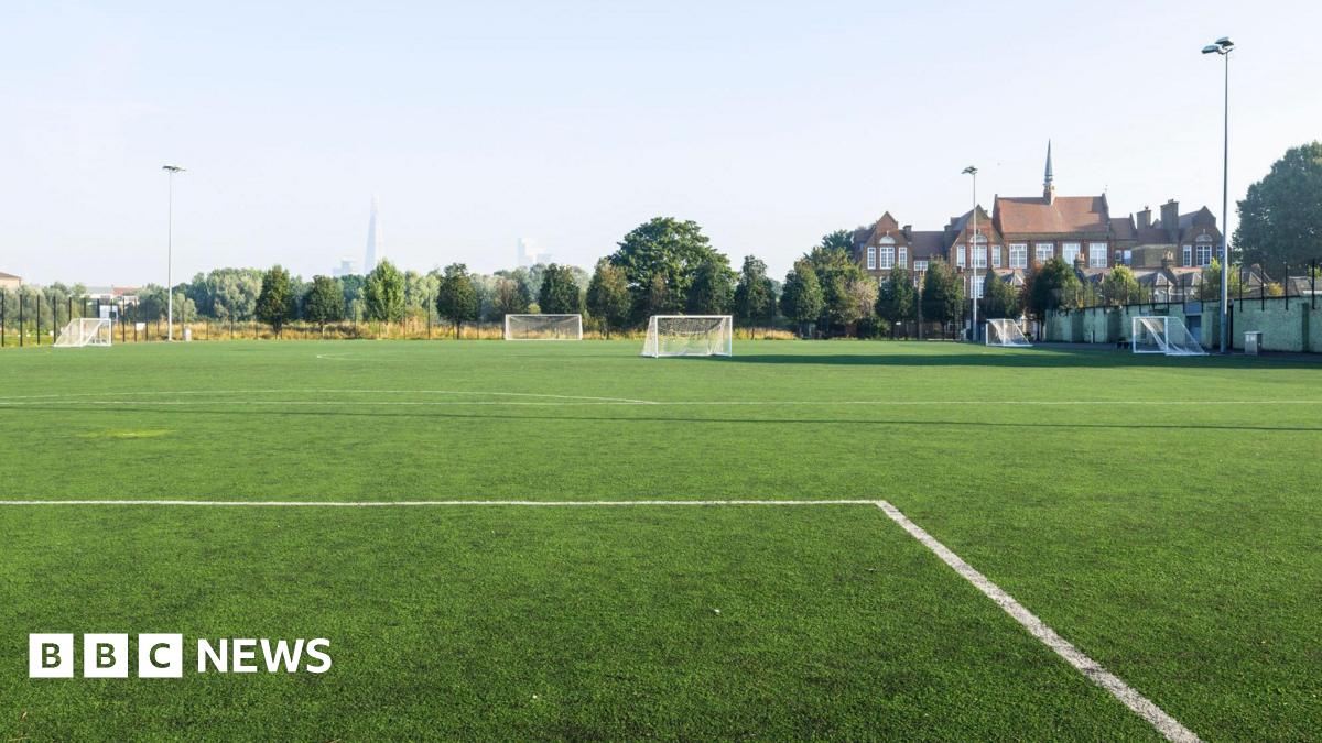 The image shows an empty leisure field in a residential district, with a view of the city in the distance. The sky is cloudless and light blue. The white lines of a football pitch can be seen in the foreground of the image.