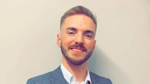Family photo Corey Longdon smiling and wearing a blue check blazer and white shirt. He has short trimmed facial hair and short dark hair