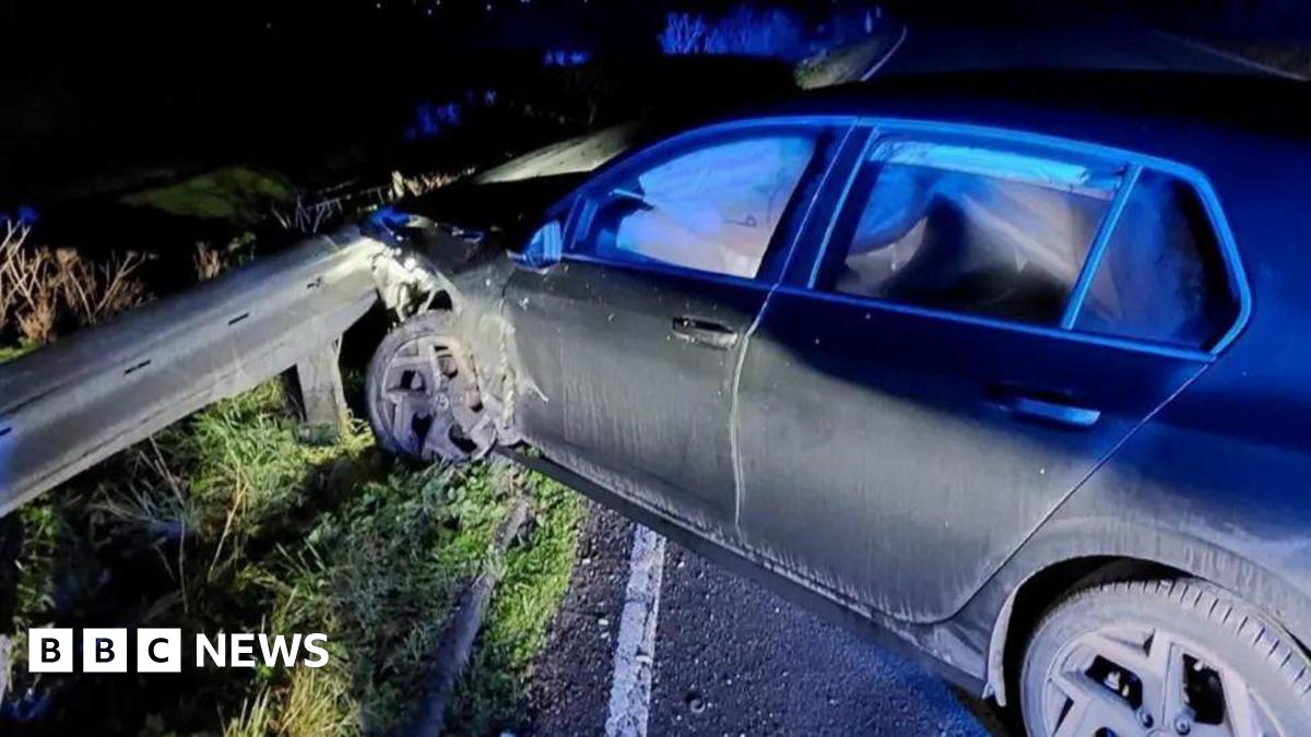 A night-time shot of a car smashed into a road barrier with the front wheel and bonnet buckled