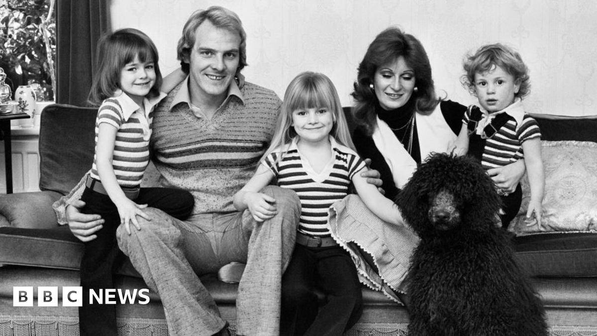 Gabby Logan and Terry Yorath at a football stadium. They are wearing formal attire with Yorath in a suit with a scarf. Logan has a jacket on with black gloves. There are people stood behind them clapping.
