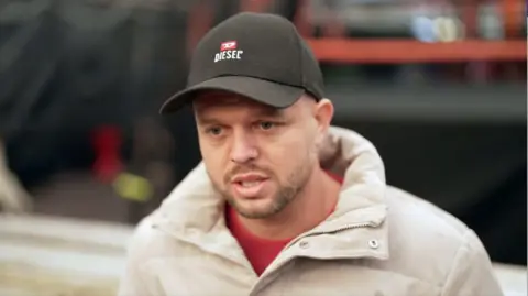 Stoner wears a light-colored padded jacket with a high collar and a red shirt underneath. He is also wearing a black baseball cap with the word “DIESEL” written on it in white letters, along with a small red logo above the text.