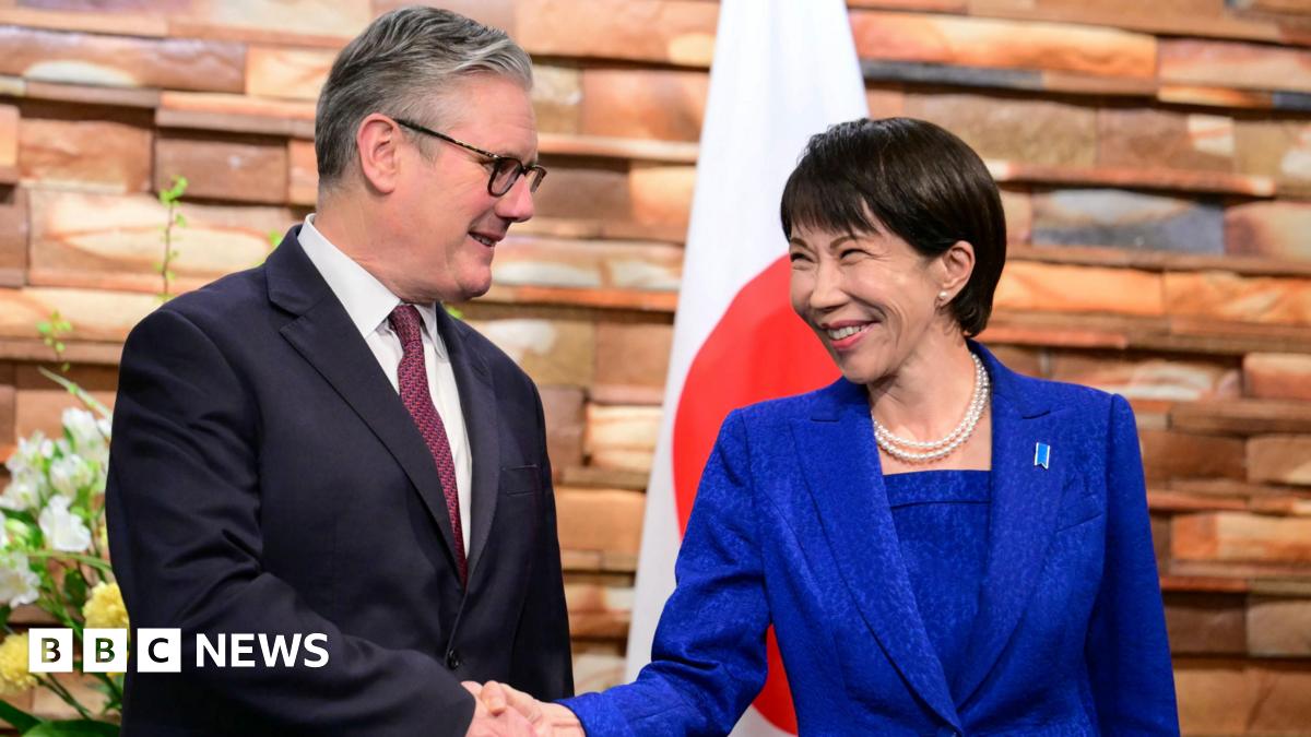 Keir Starmer and Sanae Takaichi wearing suits, shaking hands, and smiling in front of a Japanese flag and flowers