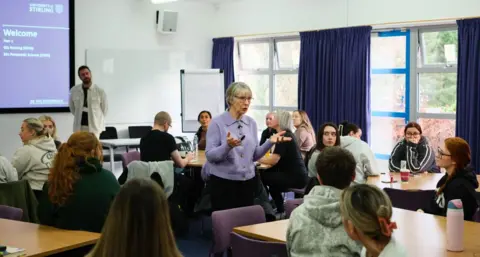 Joan Lyon, a 75-year-old woman, with grey bobbed hair and glasses, wearing a lilac cardigan lectures a room full of students