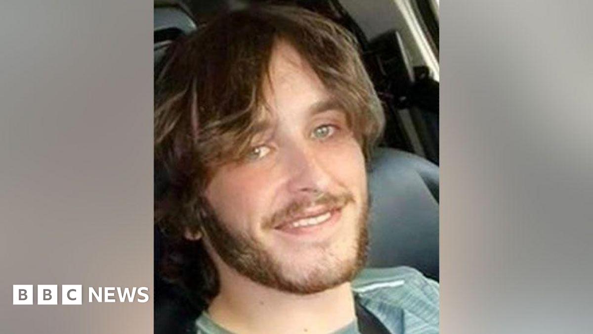 Sam Rimmer has long, light brown hair and a short beard. He has green/blue eyes and smiles for the  camera in what appears to be a selfie taken while in a vehicle.