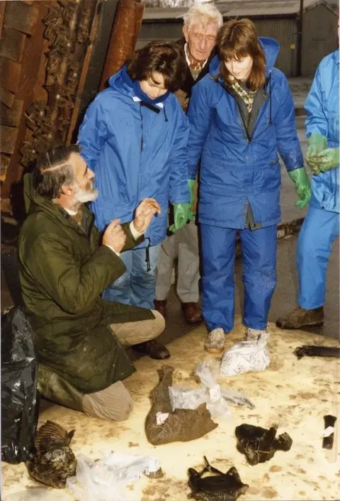 Shropshire Museums Two women with brown hair are wearing blue waterproof jackets and trousers. A man with white hair is leaning over their shoulders. Next to them is a man with brown hair and a grey beard crouching down and speaking to them. On the ground is a beige cover. There are mud covered bones on the cover
