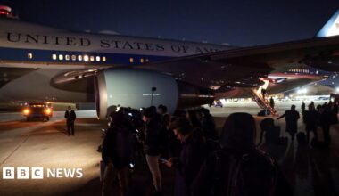 A group of journalists stand on the tarmac next to the Air Force One jet. The white and blue plane stands in the background, with the 'UNITED STATES OF AMERICA' logo visible on the side of the aircraft. The press wait in the dark, dressed in dark colours and checking their mobile phones.