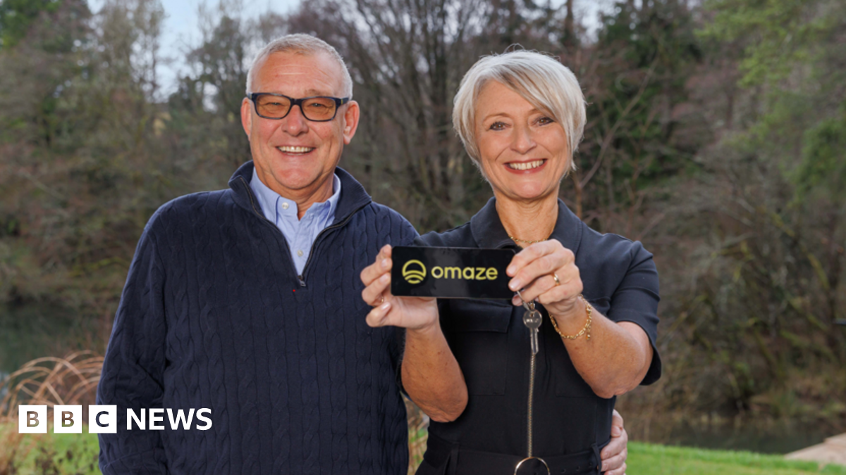 Martyn and Nicola Dickinson stand in front of a forested area. They are holding a key which says "Omaze". The man has his hand on the woman's waist and they are smiling widely.