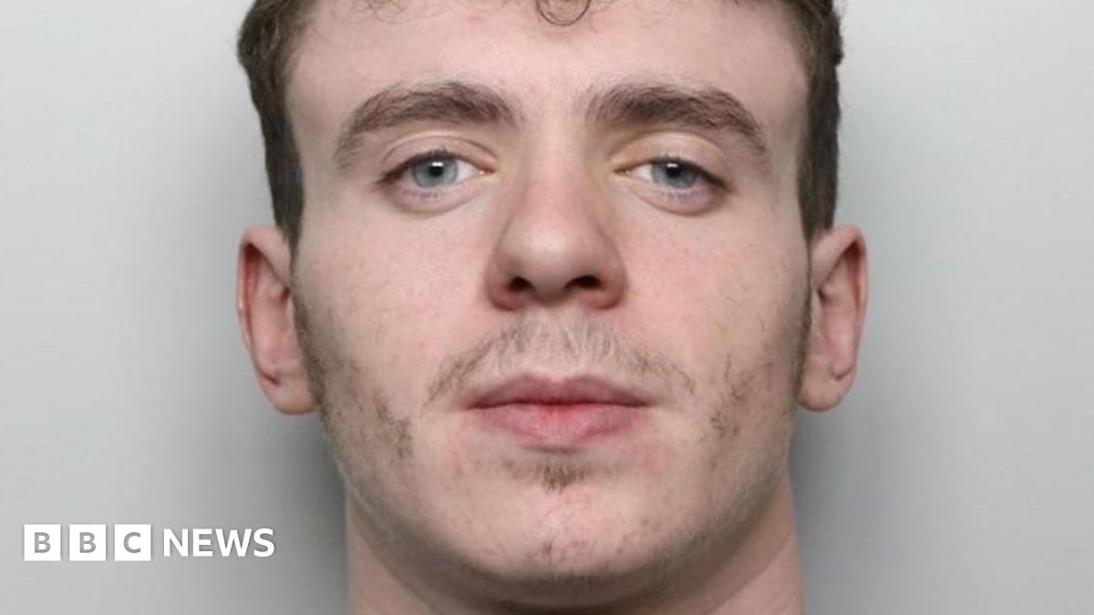 A mugshot of a 20-year-old man. He has dark brown hair which is short on the sides and curly on the top, bushy eyebrows and minimal facial hair. He appears to be smirking slightly. He is wearing a grey sweatshirt and is standing in front of a pale, plain background.