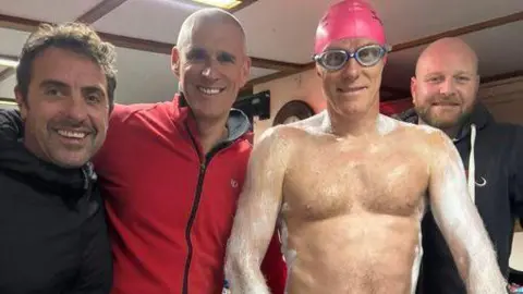 Chris Murray A man is smiling while wearing a swimming cap and goggles and what appears to be cold water surf cream on his arms and shoulders. He is standing between three other men who are also smiling.