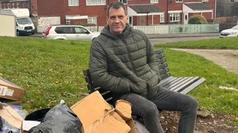 A man with a green coat sitting on a bench with recycling building up in a pile next to him. He has a slight smile. 