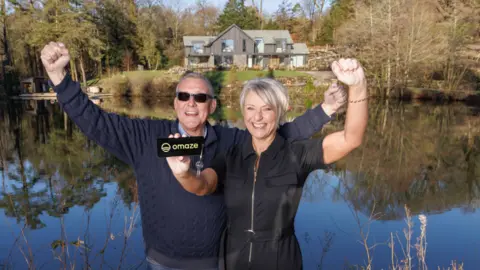 Omaze Martyn and Nicola Dickinson smile and pump their hands into the air with joy as they stand in front of a large house surrounded by water and trees.