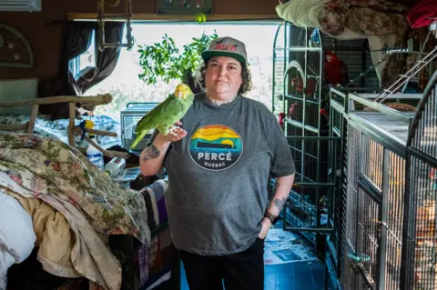 Jillian Lucas, in a grey T-shirt and baseball cap, holds a parrot on her hand at home in front of cages with other parrots. 