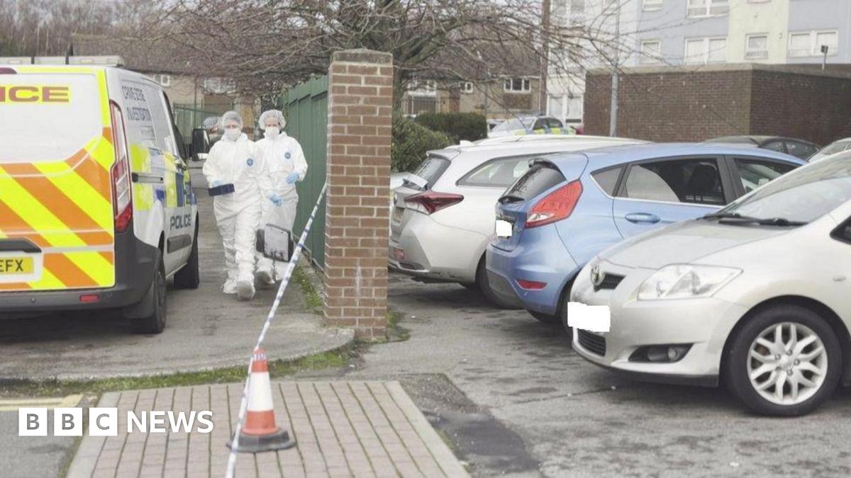 Two people wearing white forensic outfits walk near a police van and a cordon. Parked cars are also next to them by a green metal and brick fence.
