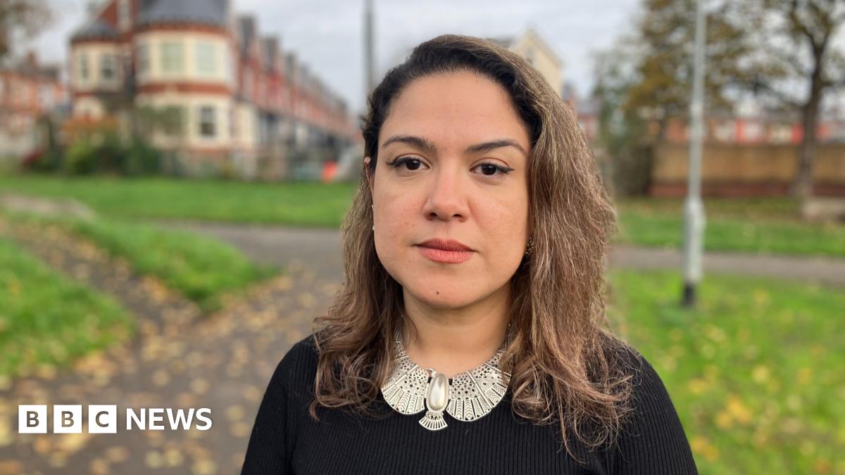 A woman with brown hair stands in a park. She is wearing a black top and large silver jewellery.