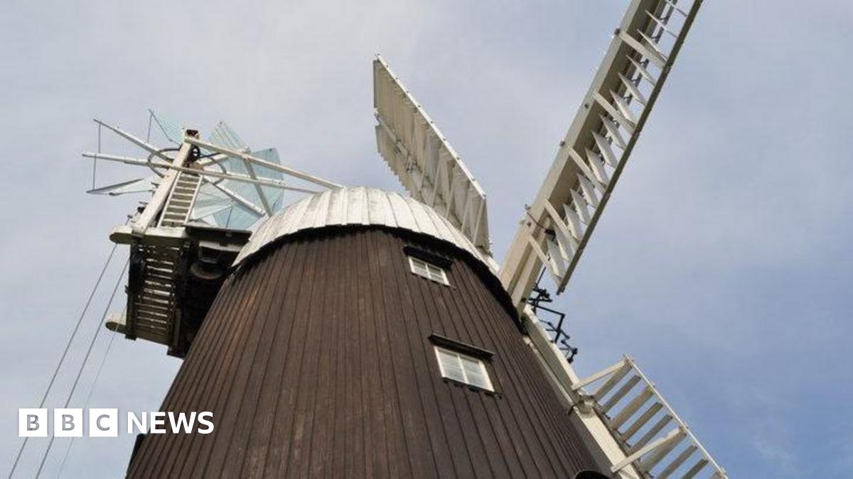 Wicken windmill in Wicken near Sohan in Cambridgeshire. The windmill has a brown exterior, reaching high into the sky. The picture has been taken from the ground and the white sails of the mill are stationery. The picture has been taken on a day with a blue sky and some overcast cloud.