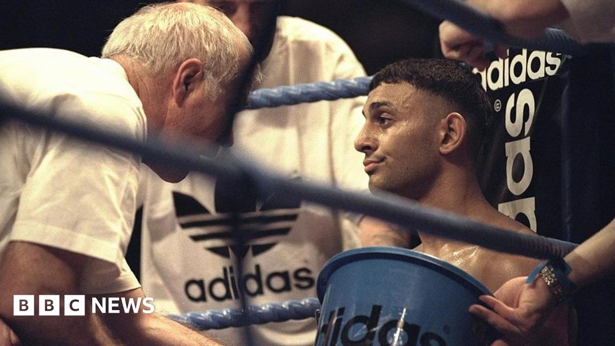 A boxer sits in the corner of a boxing ring, being attended to by his team - one holding an Adidas-branded bucket and others dressed in Adidas gear - offering support and guidance between rounds.