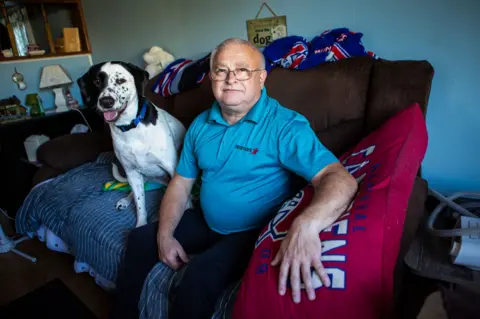 Kevin Strickland sits on his dark brown sofa in his living room next to his black-and-white dog, wearing a blue polo shirt. Behind him is a blue wall. 