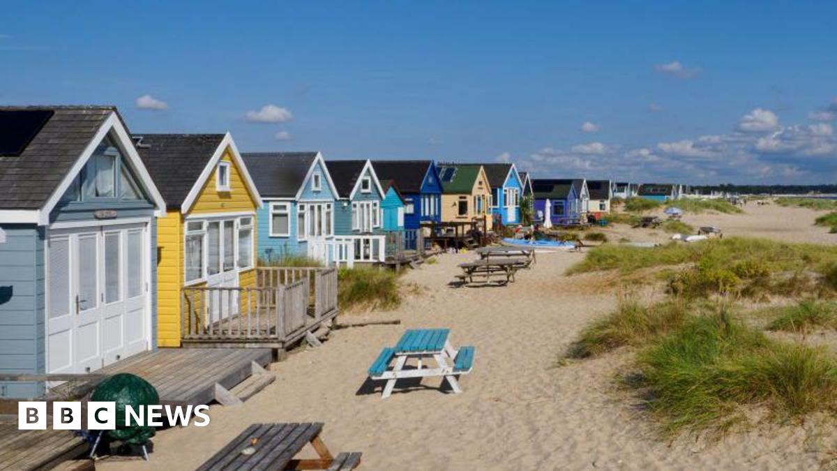 A row of brightly coloured beach huts stand on a sandy beach next to some small dunes. There are picnic benches outside the huts. The sky is blue and it is a sunny day.