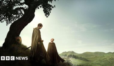 Publicity shot from A Knight of the Seven Kingdoms. Shows Ser Duncan the Tall and Egg standing beside a curved tree with mountains in the background. Ser Duncan is wearing a light coloured cloak, has dark brown hair and carrying a sword looking down at Egg. Egg is a young child and is bald, wearing a light coloured cloak and looking up at Ser Duncan.