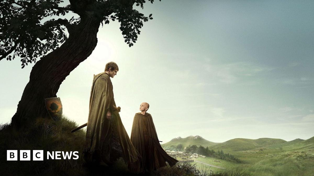 Publicity shot from A Knight of the Seven Kingdoms. Shows Ser Duncan the Tall and Egg standing beside a curved tree with mountains in the background. Ser Duncan is wearing a light coloured cloak, has dark brown hair and carrying a sword looking down at Egg. Egg is a young child and is bald, wearing a light coloured cloak and looking up at Ser Duncan.
