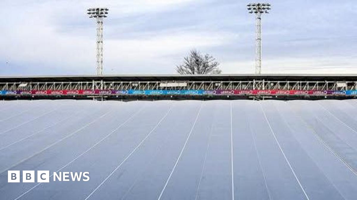 A covering over the top of Luton Town Football Club's pitch, it is white and very large with lines across it. A football stand can be seen in the distance, with a free behind it and two large flood lights.