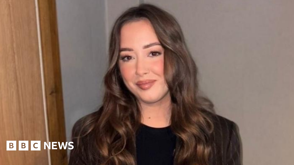 Laura is a young woman with long, brown hair. She is smiling at the camera. She is standing against a white wall and is wearing a brown pinstripe blazer and a black t-shirt.