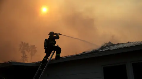 Justin Sullivan / Getty Images A firefighter, dressed in protective clothing, is silhouetted as they fire a water cannon at a burning house with the sun shining through a hazy sky in the background, in Altadena on 9 January 2025. 