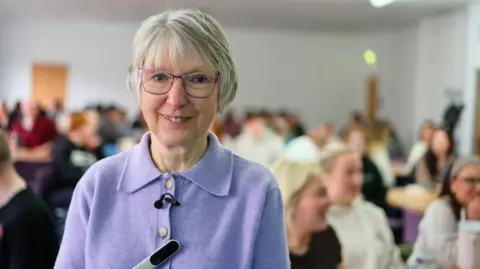 BBC Joan Lyon, a 75-year-old woman, with grey bobbed hair and glasses, wearing a lilac cardigan. Behind her are groups of students sitting at tables..